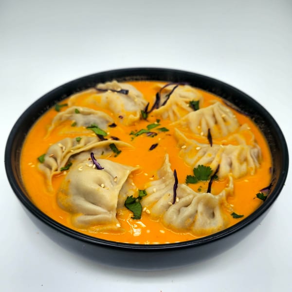 Nepalese momos in a vibrant orange sauce, garnished with sesame seeds and cilantro in a black bowl.