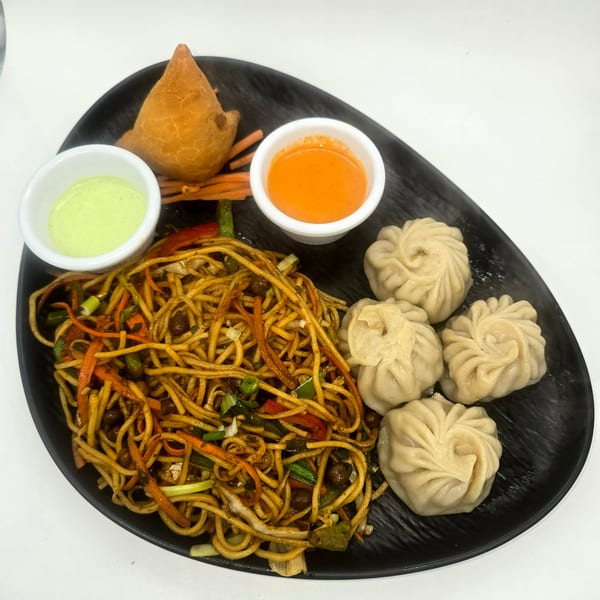 A black plate with vegetable chowmein, momos, a samosa, and two dipping sauces.
