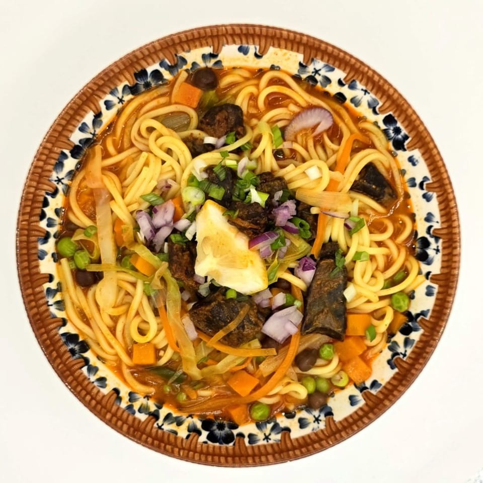 A close-up of a bowl of Nepalese thukpa noodles with vegetables, meat, and a lemon wedge.